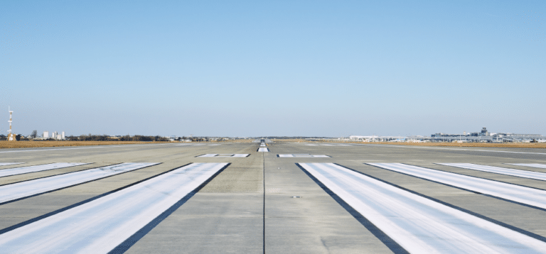 View down the center of a wide airport runway with clear blue sky, white markings, and airport buildings—expertly maintained thanks to professional Commercial Restoration Services—visible in the distance.