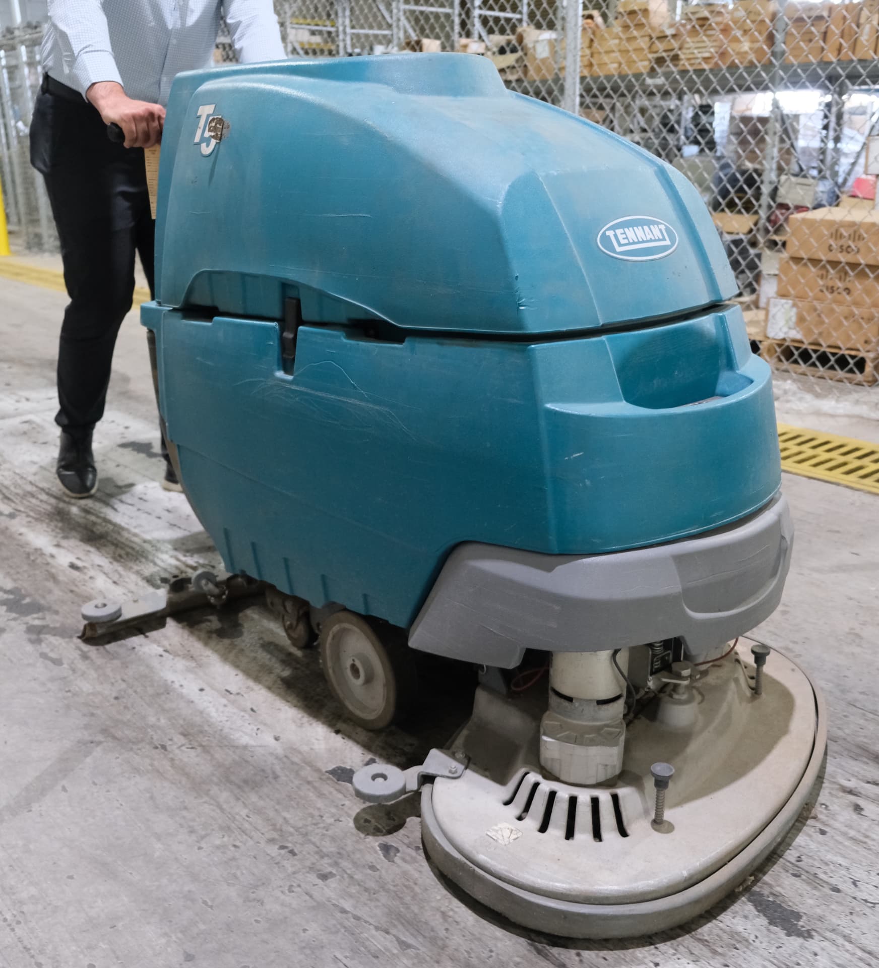 A person operates a large blue Tennant floor scrubber on a warehouse floor near a chain-link fence and shelves filled with boxes, maintaining the facility as part of comprehensive Commercial Restoration Services.
