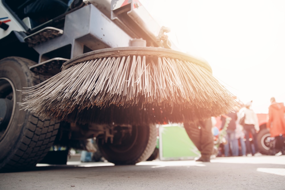 Close-up view of a street sweeper brush attached to a vehicle, with people and sunlight visible in the background—an essential part of maintaining clean urban spaces, much like Commercial Restoration Services support the upkeep and renewal of city environments.