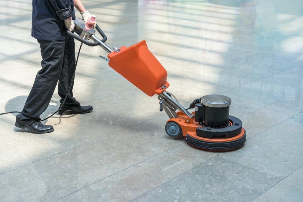 A professional from Commercial Restoration Services is operating an orange floor polishing machine on a large, shiny tiled surface in a brightly lit indoor area.