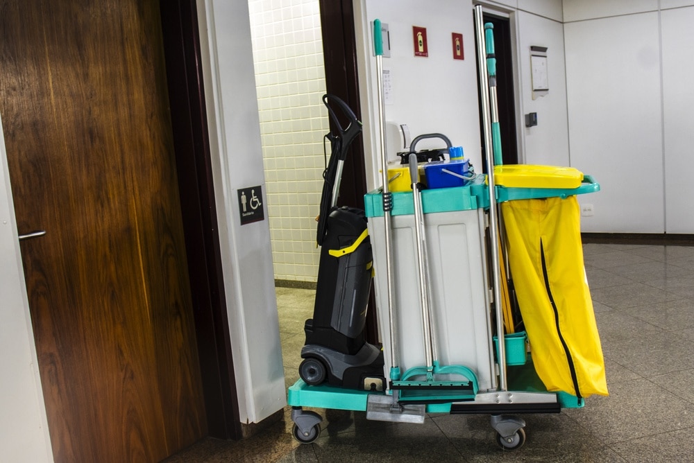 A janitorial cart with cleaning supplies and a yellow bag, often seen as part of Commercial Restoration Services, is parked in a hallway outside a restroom door.