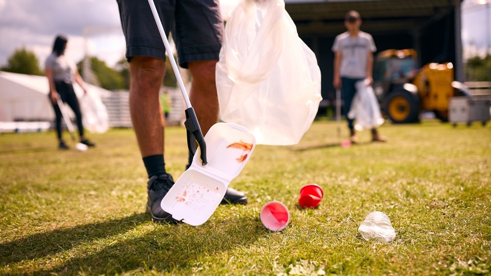 Person using a litter picker to collect trash, including a white food container and red cups, on a grassy field with others cleaning in the background—demonstrating community care that reflects the thorough standards of Commercial Restoration Services.