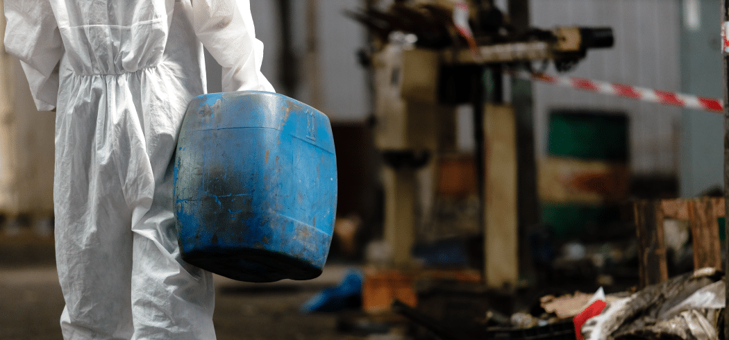 Person in protective suit carrying a blue plastic container in an industrial setting with scattered debris and caution tape in the background, exemplifying the professionalism and safety measures of Commercial Restoration Services.