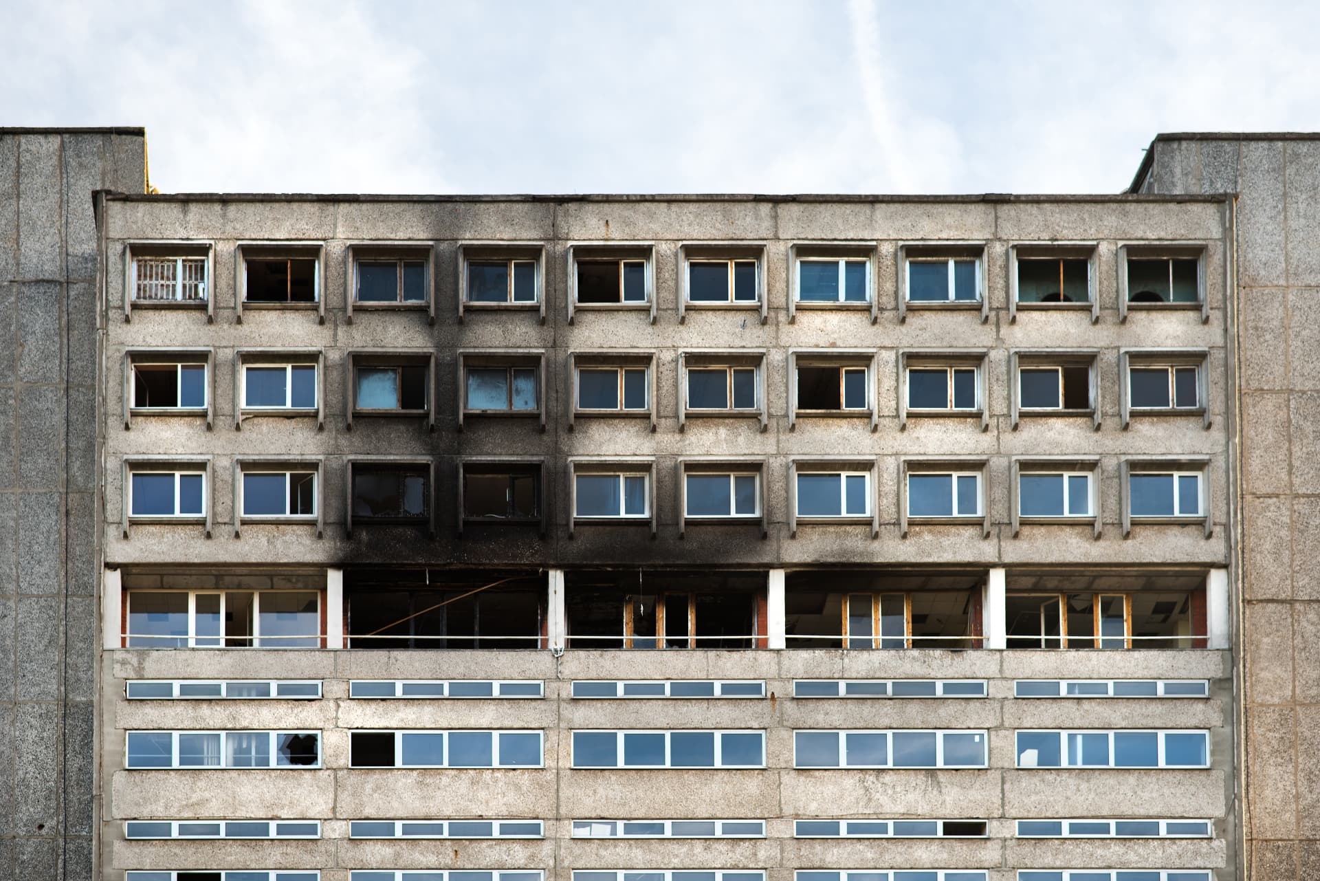 Gray concrete apartment building facade with visible fire damage on the upper floors; some windows are missing or broken, others are intact. The sky is visible in the background, highlighting the urgent need for professional Commercial Restoration Services to restore safety and functionality to the property.