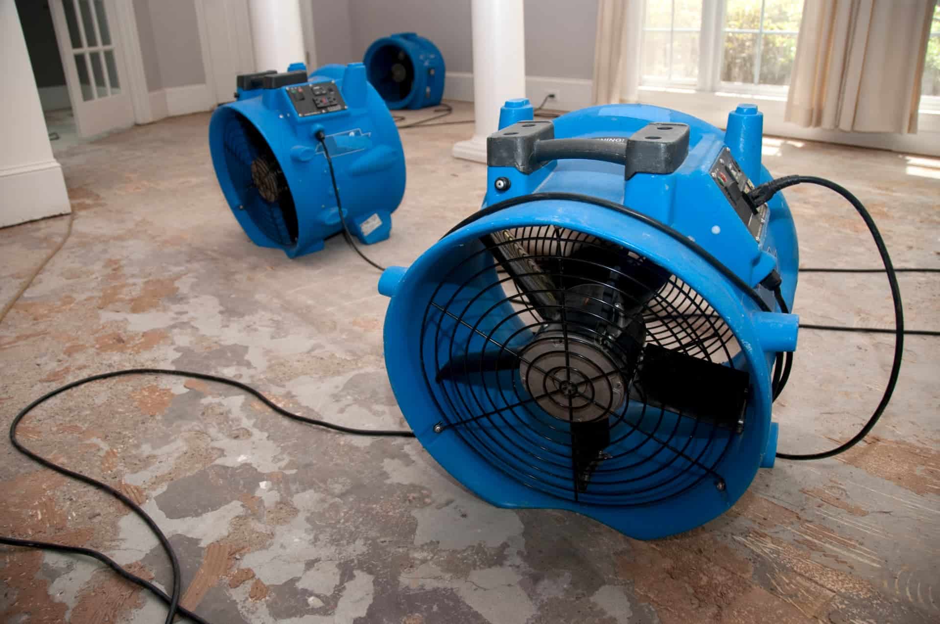 Three blue industrial floor dryers are positioned on a bare, damaged floor in a room with large windows and white columns. Electrical cords are visible, highlighting the ongoing work by Commercial Restoration Services to restore the space efficiently.