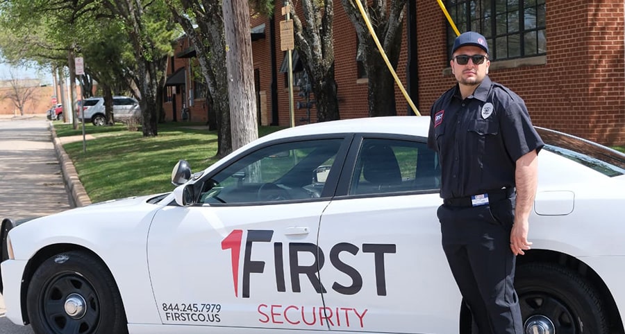 A security guard in uniform stands next to a white First Security patrol car parked by a red brick building on a sunny day, ensuring the safety of the premises as Commercial Restoration Services are underway.