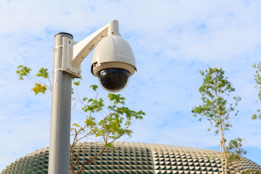 White security camera on a tall pole with trees and a domed building in the background under a blue sky, highlighting the importance of vigilant monitoring for sites undergoing Commercial Restoration Services.