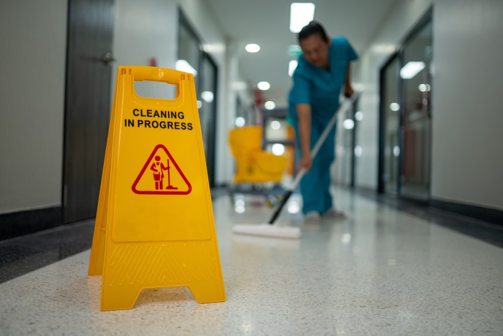 A yellow “Cleaning in Progress” sign stands in a hallway as a person in blue scrubs mops the floor in the background, exemplifying the attention to detail provided by professional Commercial Restoration Services.