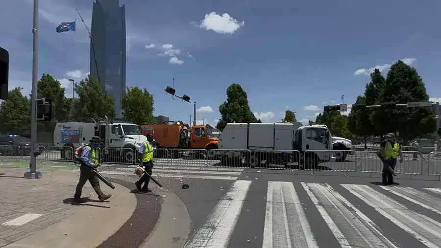 City workers clean debris from a crosswalk with leaf blowers near several sanitation trucks, while Commercial Restoration Services crews stand by to assist. Tall buildings and trees rise in the background under a blue sky.