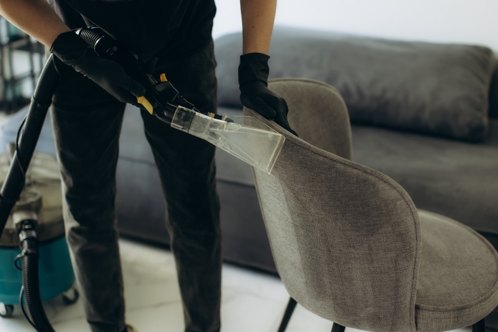 A person wearing gloves uses a vacuum cleaner with a special attachment to clean the fabric of a gray upholstered chair, demonstrating the meticulous care provided by Commercial Restoration Services. A gray sofa and a cleaning machine are visible in the background.